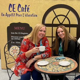 Two Salus faculty members sitting at a table, one holding a tea cup with a sign that says "Ce Cafe, Un Appetit Pour L'education" behind them 