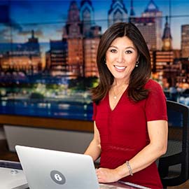 Photo of Nydia Han sitting at 6ABC Action News Desk in front of computer with city of Philadelphia in background