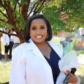 Makayla Cain Standing outside of the Hafter Center wearing her white coat and holding a bouquet of flowers in her hand 