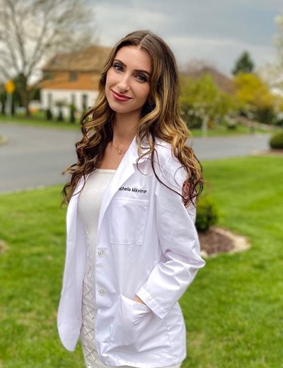 Michelle standing outside smiling at the camera wearing a white coat and white dress