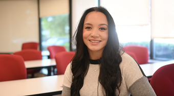 Student Emaree Stone shown in white shirt in a well lit Salus clasroom with red chairs, white tables, and windows in the background.