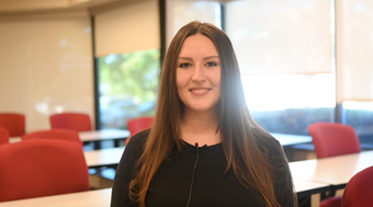 Student Nicole Jeziorski shown in black shirt sitting in a well lit Salus classroom with red chairs, white tables, and windows in the background.