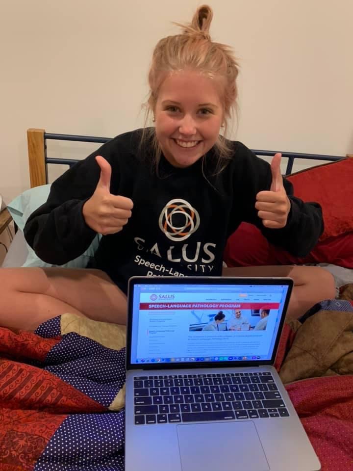 Rachel sitting on her bed with her laptop wearing a Salus University sweatshirt