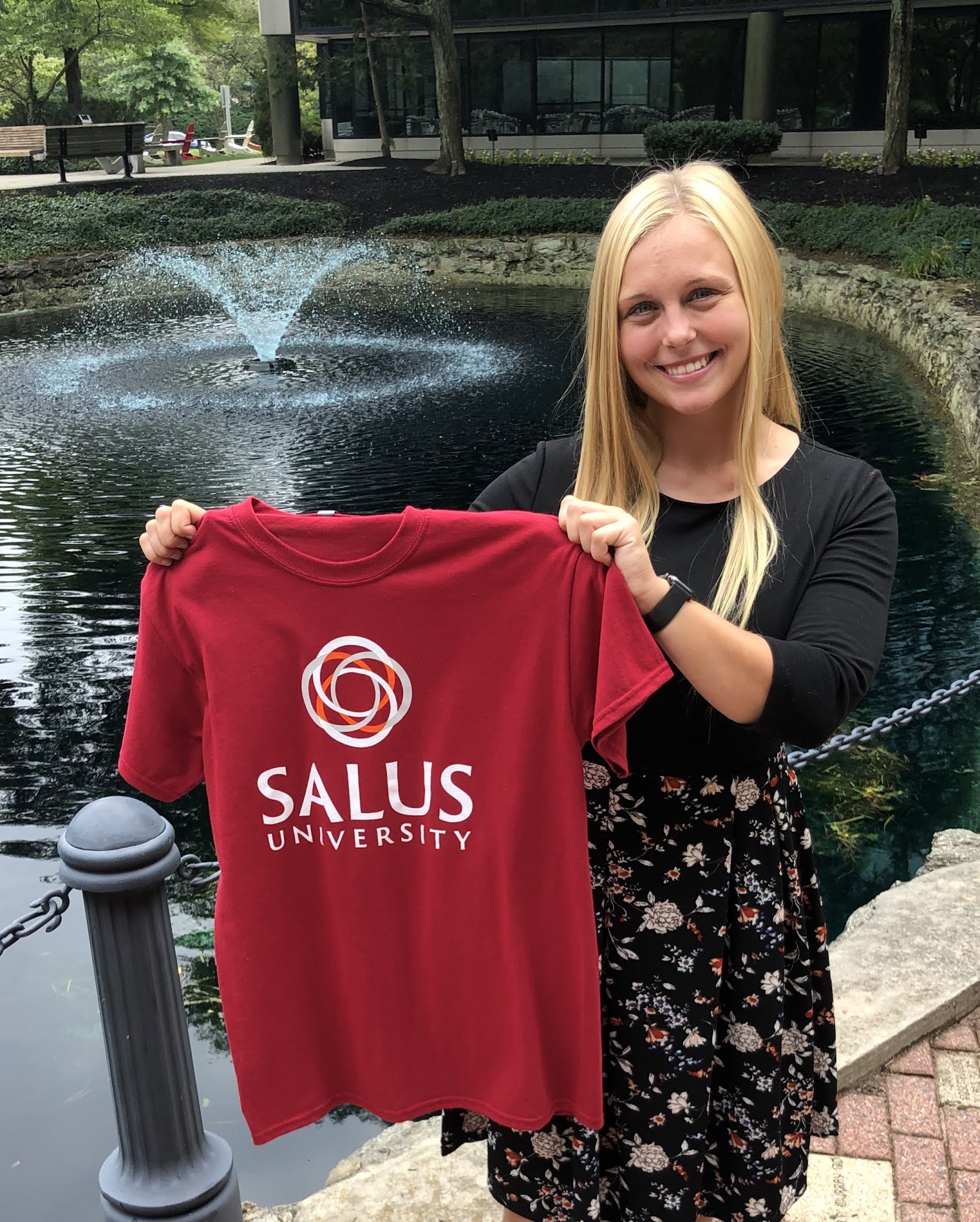 Robin holding a Salus University t-shirt in front of the Salus pond